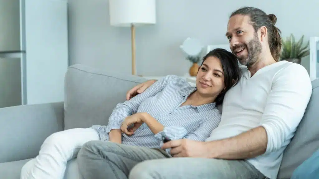 A man and woman sit closely on a grey sofa while looking toward the side.