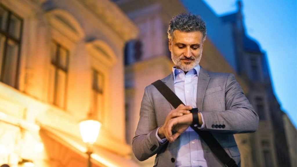 A man in a grey suit looks at his watch while standing on a city street.