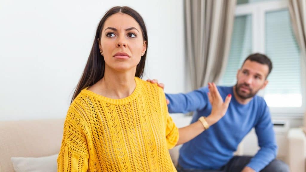 A woman in a yellow sweater gestures with her hand while a man sits blurred behind her.