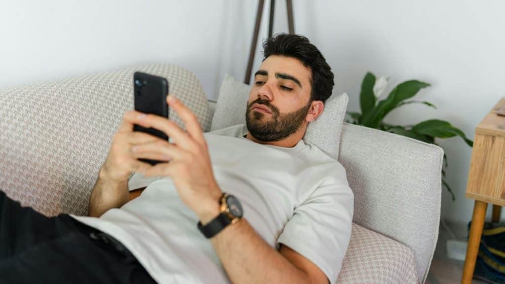 A man reclines on a couch while looking down at a smartphone in his hands.