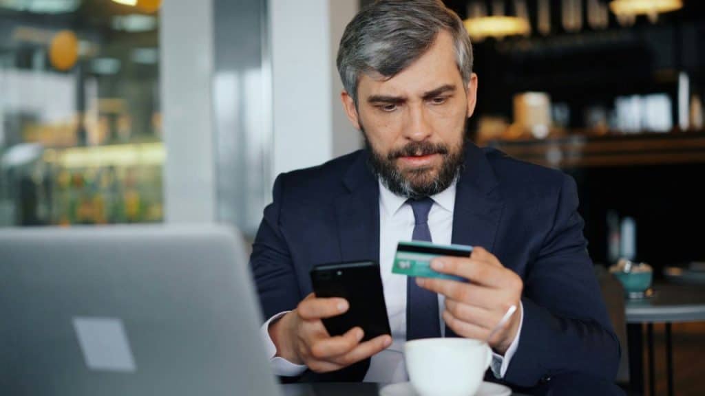 A man in a suit looks at a smartphone while holding a credit card in a cafe.