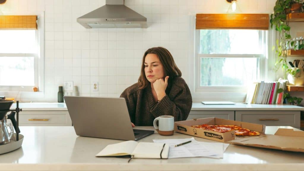 A person sitting at a kitchen counter working on a laptop beside papers and a pizza box.