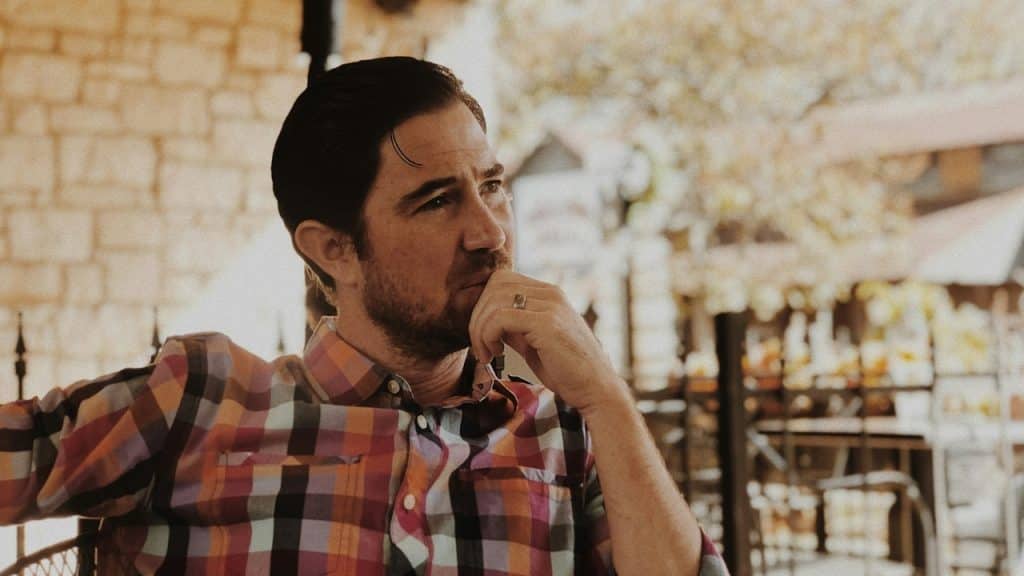 A man in a colorful plaid shirt sits outdoors, resting his chin on his hand.