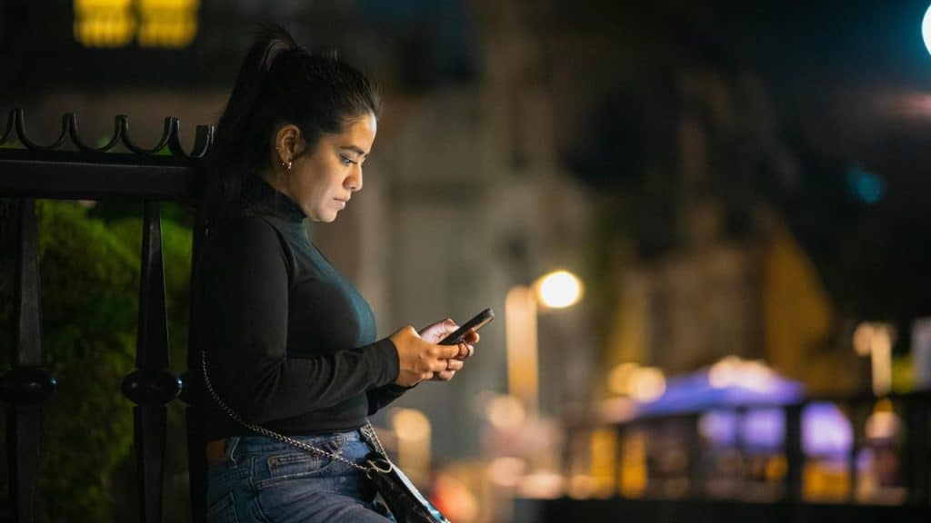 A person standing outdoors at night, focused on a phone under city lights.