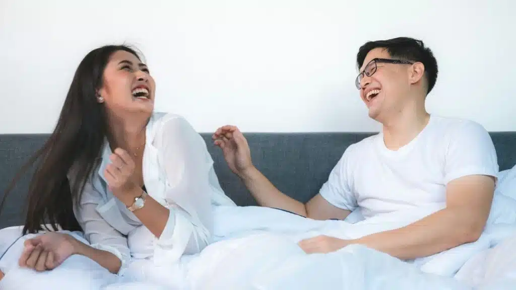 A man and woman in white shirts laugh together while sitting on a bed.