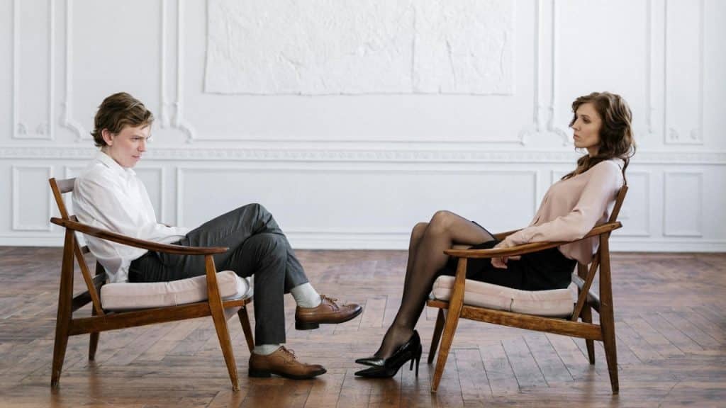 A man and woman sit in wooden chairs facing each other in a large, empty room