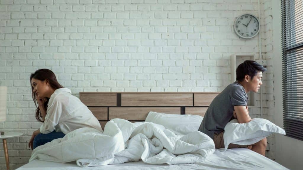 A man and woman sit on opposite sides of a bed looking away from each other.