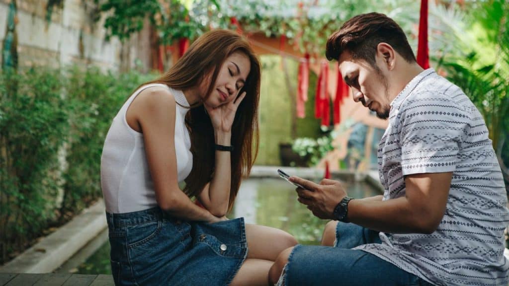 A woman rests her head on her hand while a man beside her looks at a phone.