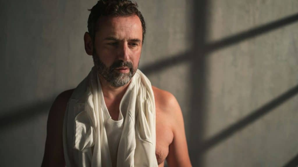 A shirtless man with a white shirt around his neck looks downward in dim light.