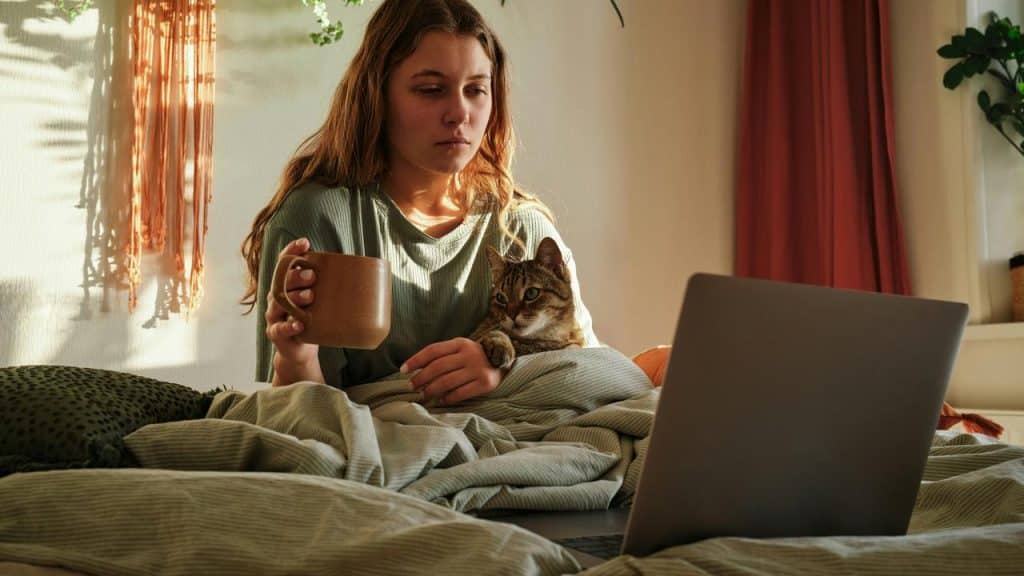 A person sitting on a bed holding a mug, with a cat on their lap and a laptop nearby.