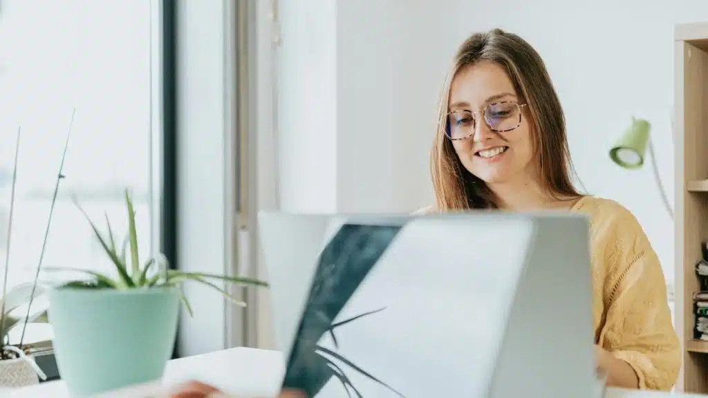 A woman smiling while working on a laptop at a bright desk.