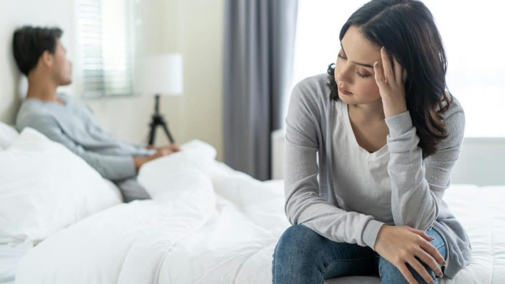 A woman sitting on a bed holding her head while a man sits behind her.