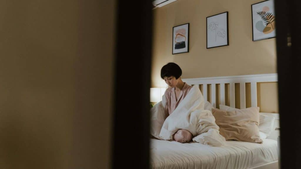 A woman sits upright in a bed, partially covered by a white duvet, looking downward.