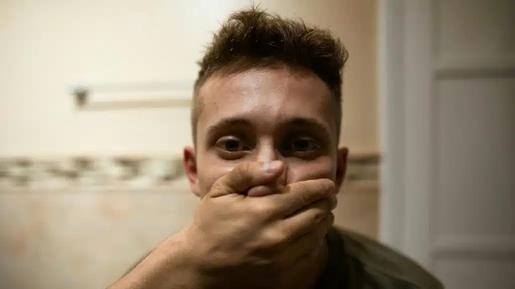 A man with brown hair covers his mouth with his hand while looking at camera.
