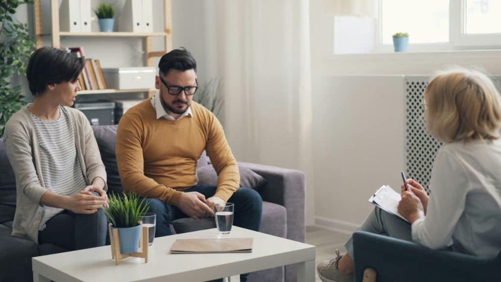 Three people sit in a room during a meeting; one person is taking written notes.
