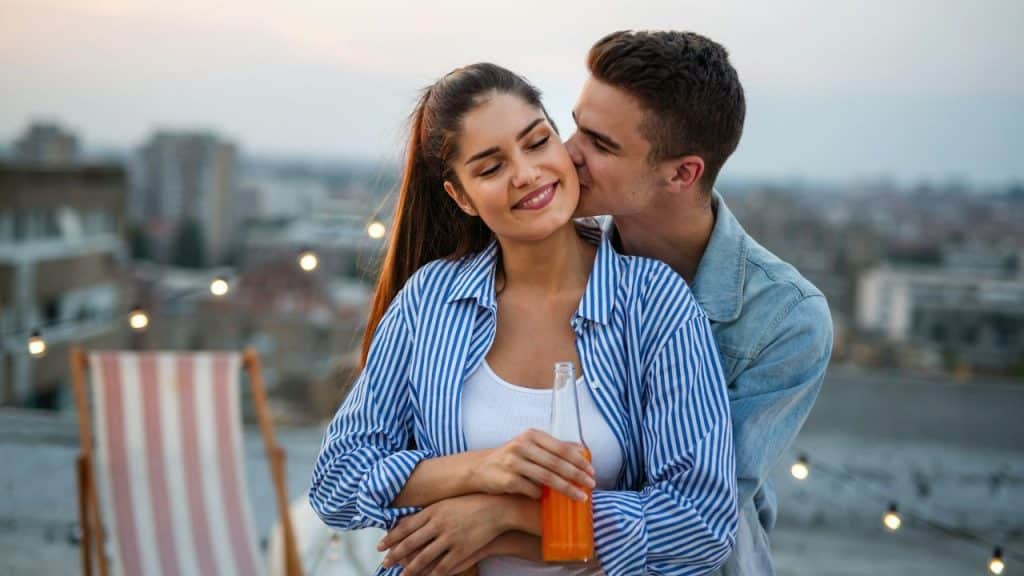 A man in a denim jacket kisses a smiling woman's cheek on a rooftop at dusk.