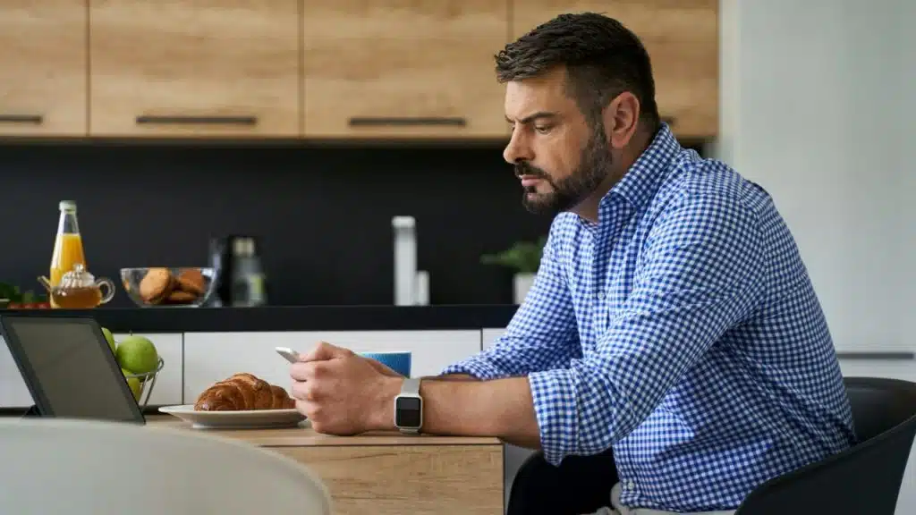 A man sits at a table with breakfast, looking at a phone and a tablet.