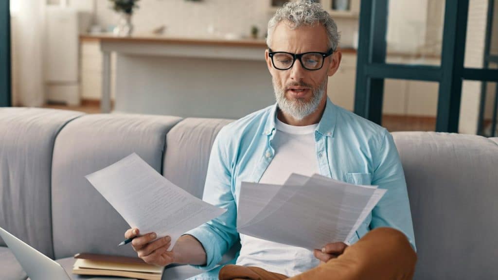 A man with a grey beard wears glasses and looks at papers in his hands.