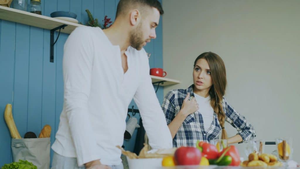 A woman in a plaid shirt gestures toward herself while speaking to a man in a kitchen.