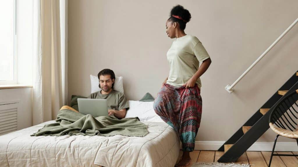 A woman stands near a bed where a man sits under blankets using a laptop.