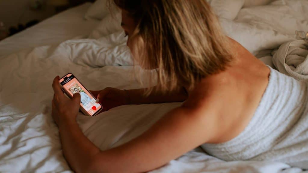 A woman lying on a bed uses a smartphone displaying a dating app profile.