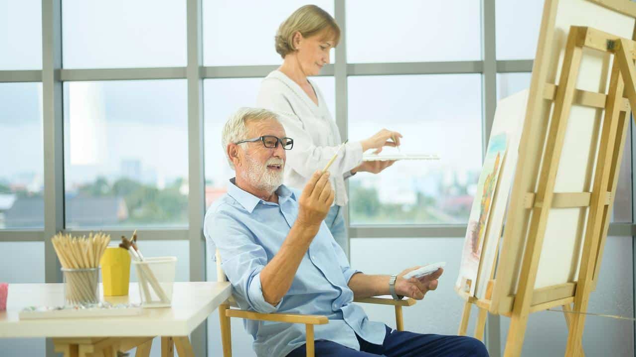 An elderly man holding a paintbrush in a bright art studio with a woman mixing paint behind him.