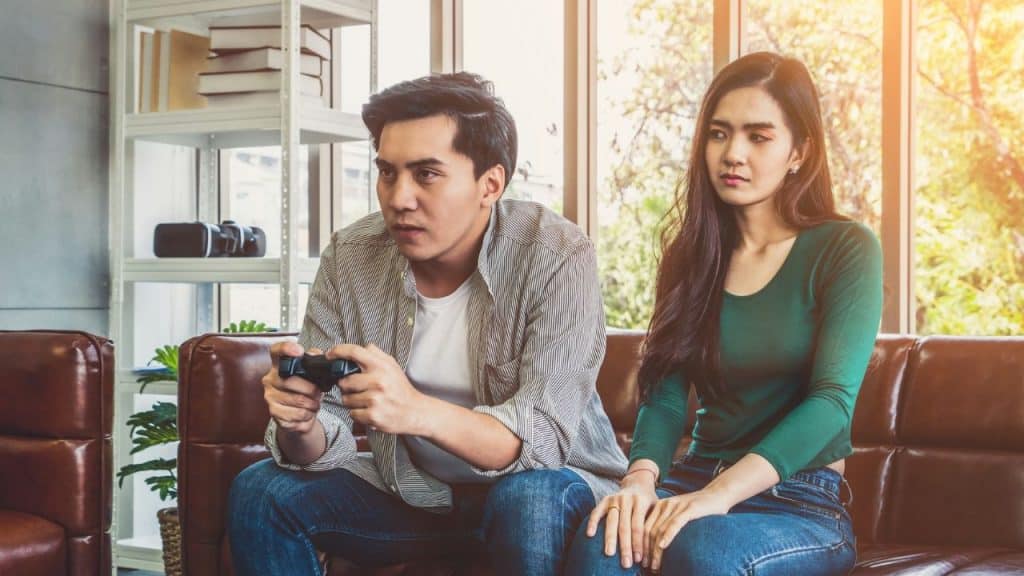 A man plays a video game while a woman sitting next to him looks away unhappily.