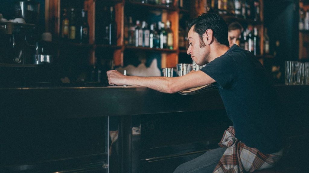 A man sits alone at a dark bar leaning forward on his elbows near rows of bottles.