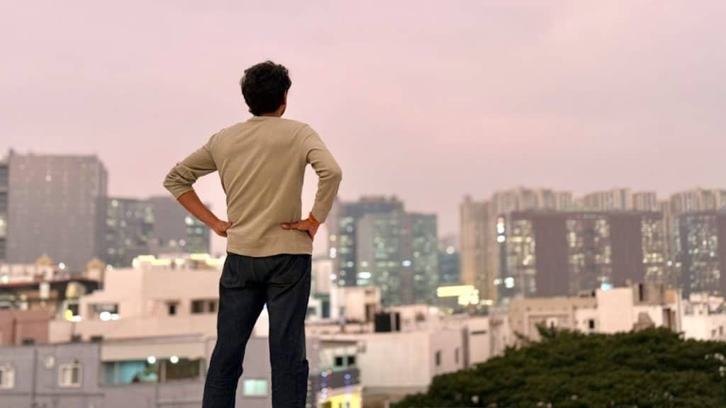A man stands on a rooftop with hands on hips, looking out at a city skyline.