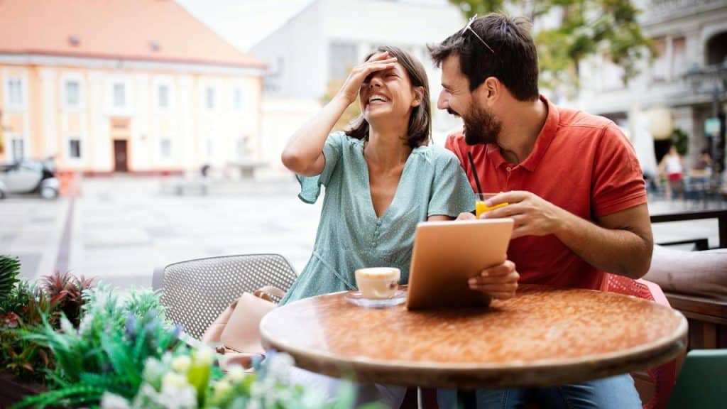 A couple laughing together while sharing a drink and a tablet at an outdoor café.