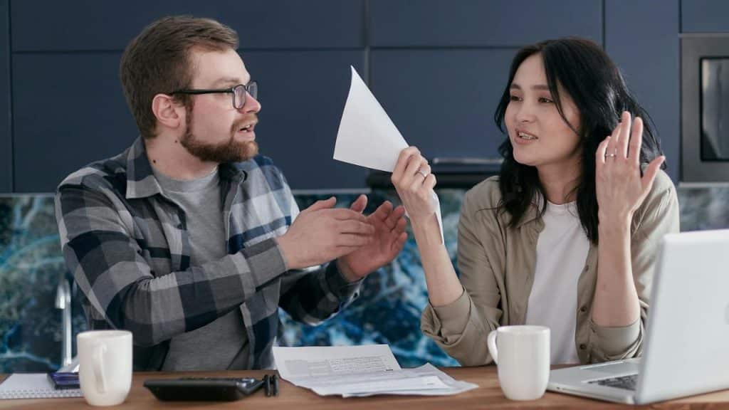 A man and woman sit at a desk with a laptop and papers while gesturing.