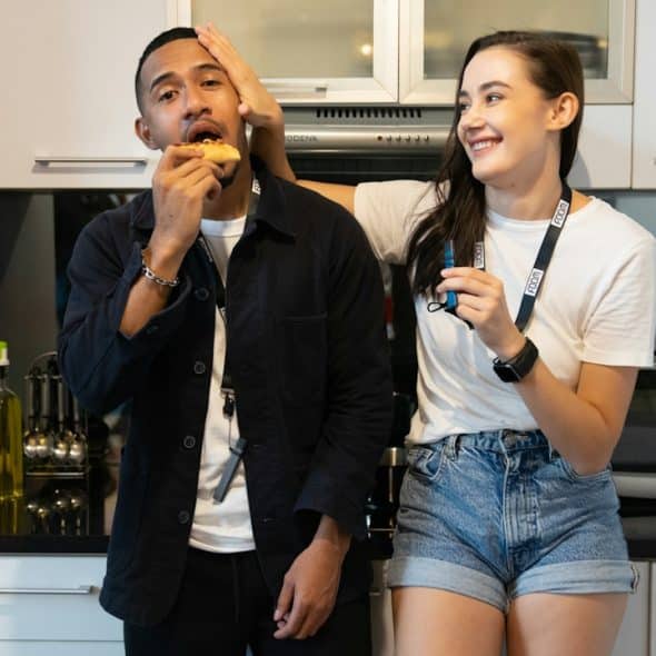 A woman playfully pushing a man’s head while he eats a slice of pizza as they lean against a cabinet in a kitchen.