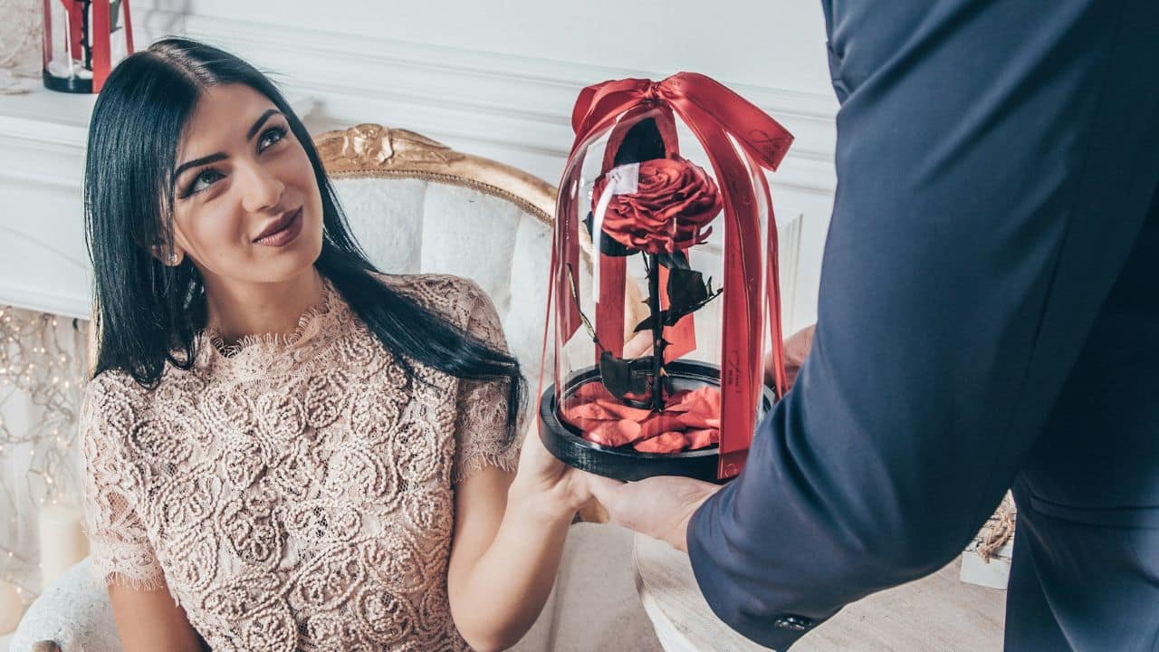 A man presents a single red rose inside a glass cloche to a seated woman.