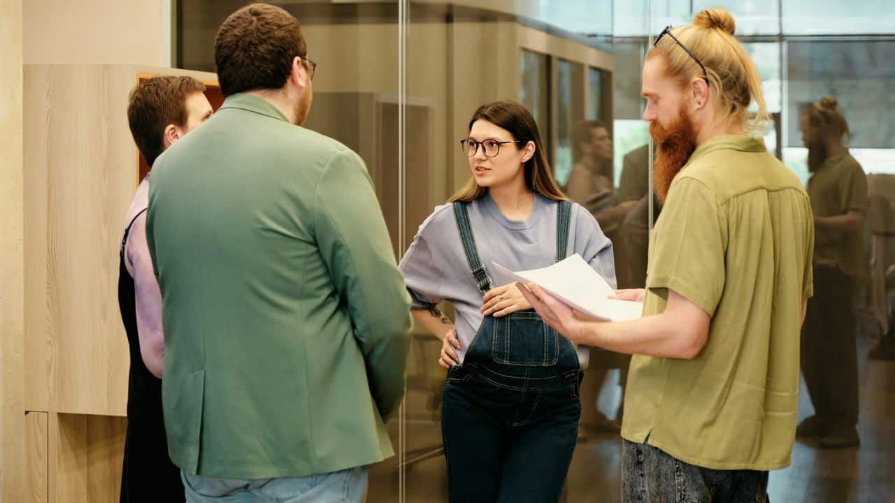 A woman wearing glasses and denim overalls stands in a group of people in an office.
