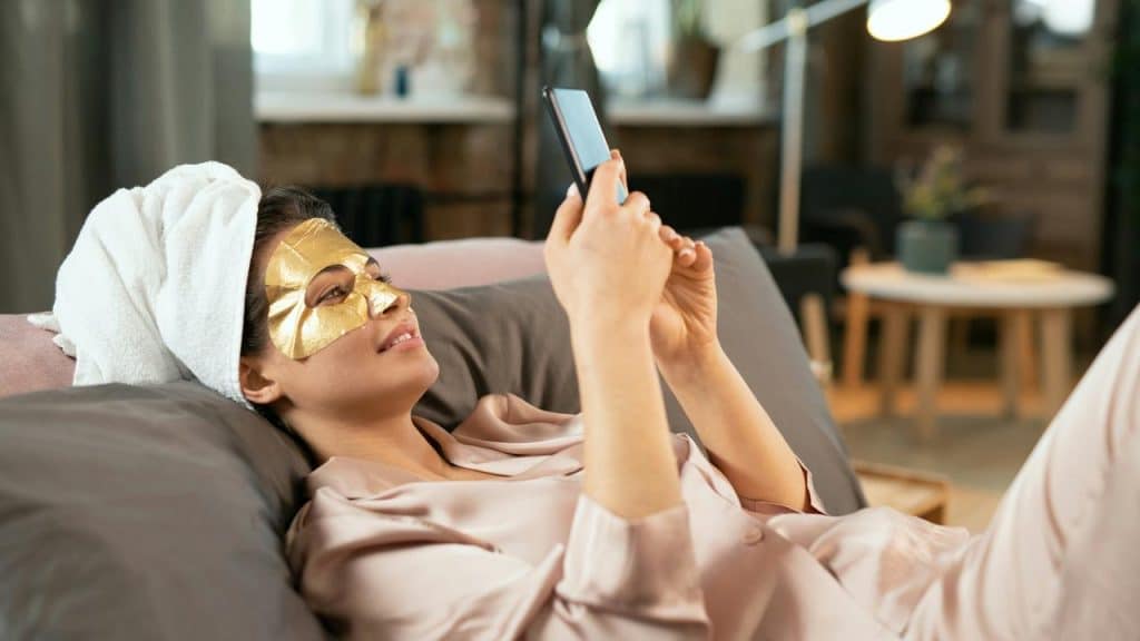 A woman lying on a couch wearing a face mask and looking at her smartphone.