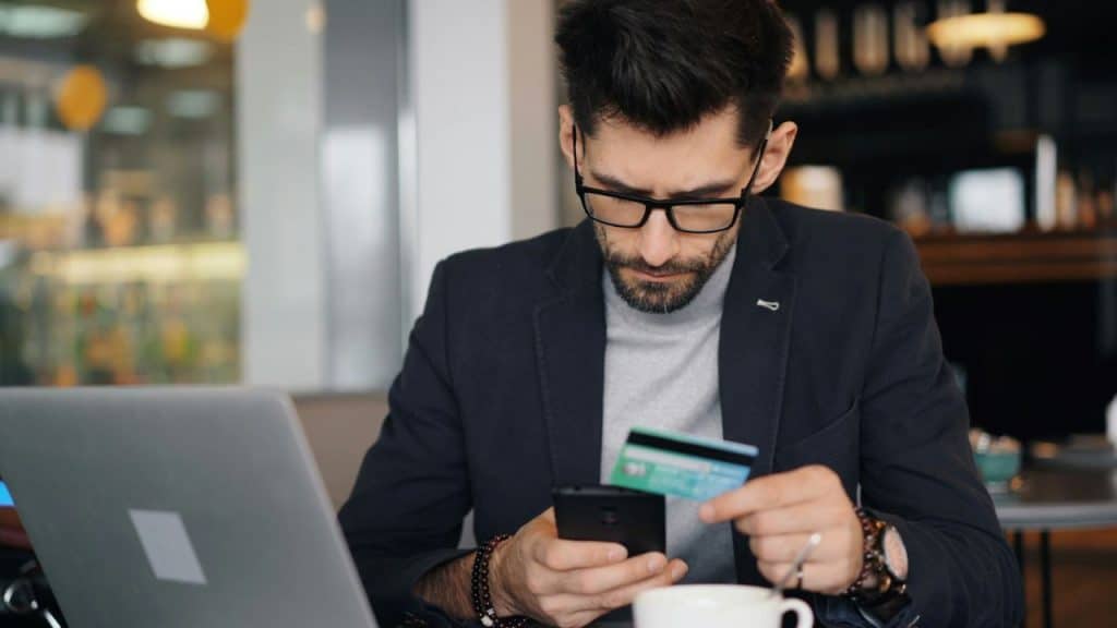 A man wearing glasses holds a credit card while looking at his smartphone near a laptop.