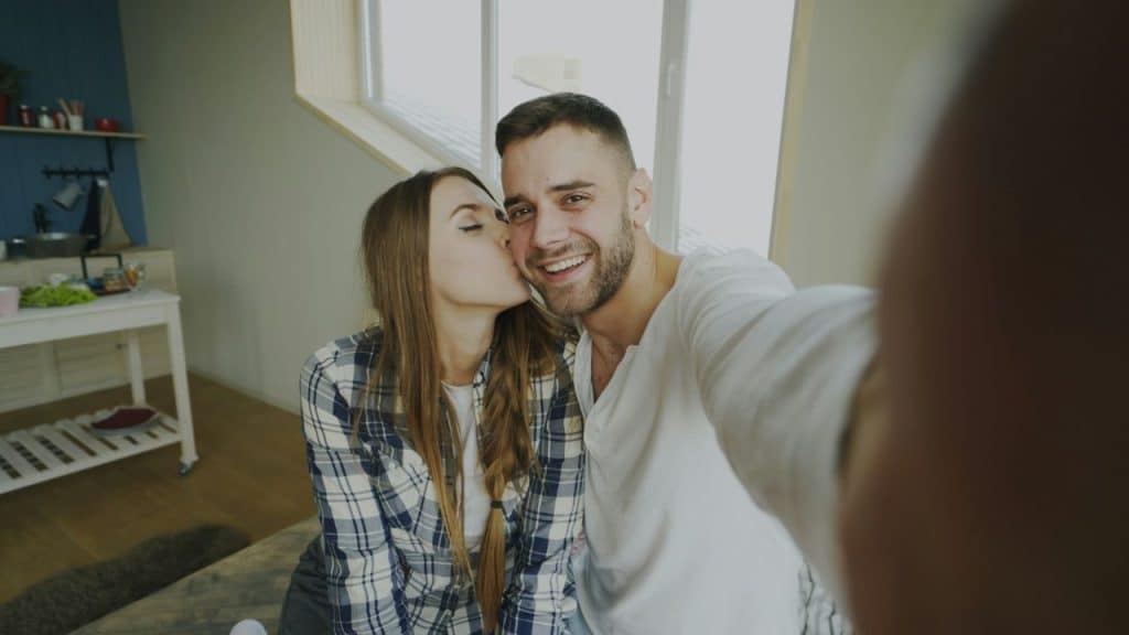 A woman in a plaid shirt kisses a smiling man's cheek while taking a selfie.