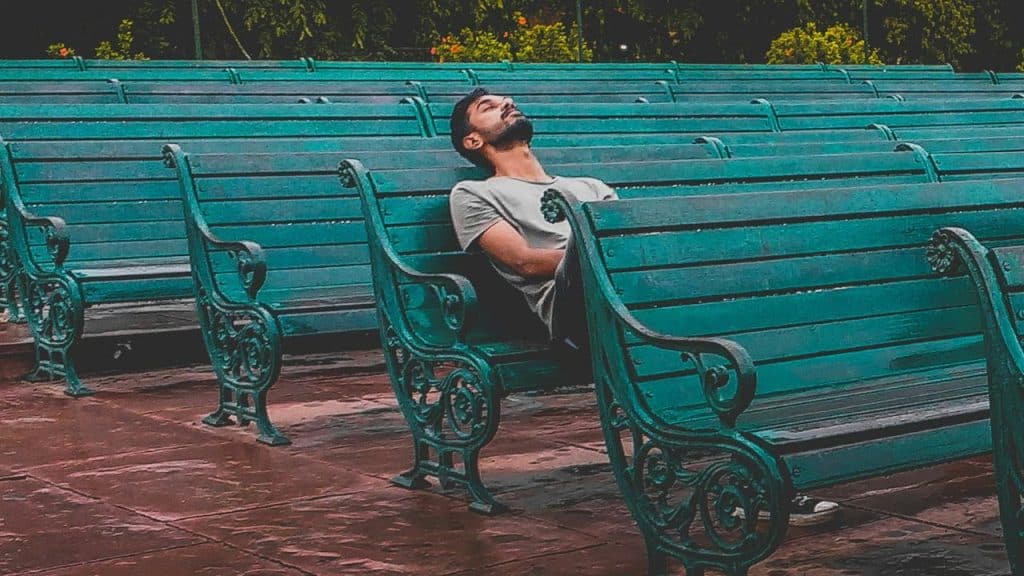 A man leans back on a green park bench with his eyes closed and arms crossed.