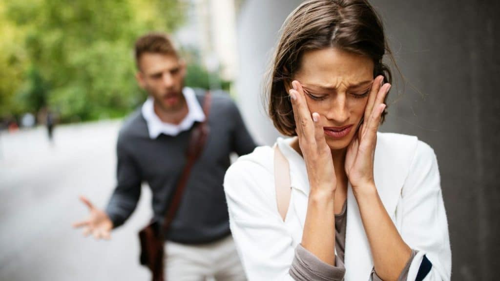 A woman covers her face with her hands while a man gestures in the blurred background.