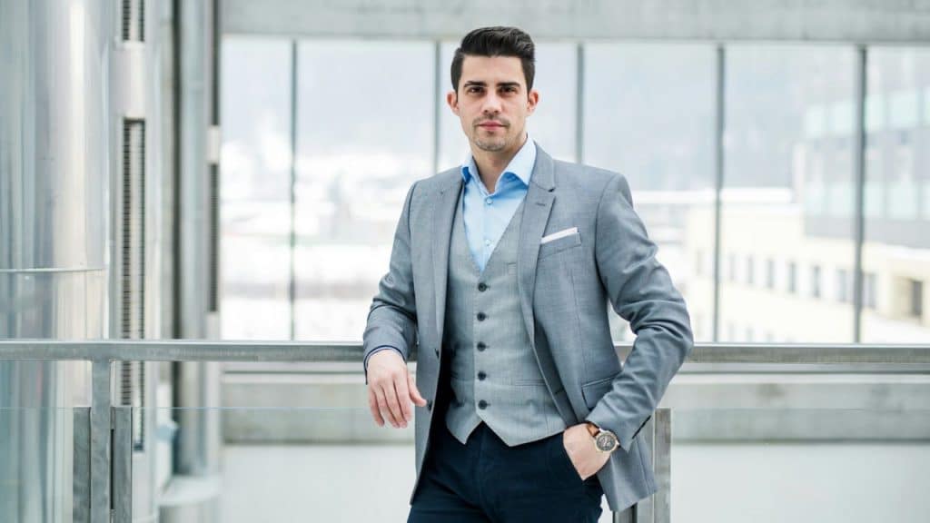 A man wearing a grey three-piece suit leans against a metal railing in a building.