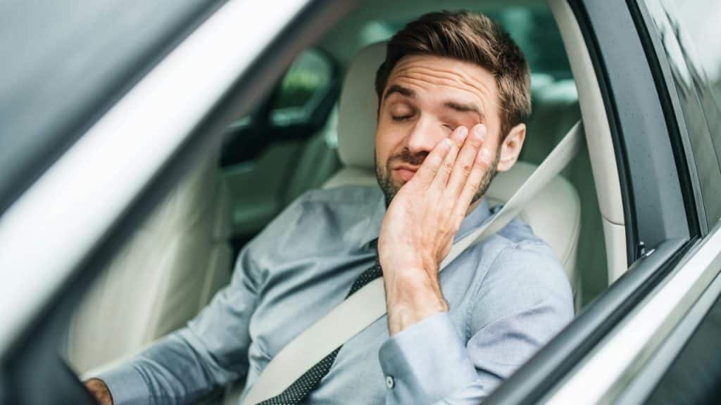 A tired man rubbing his eye while sitting in the driver’s seat of a car.