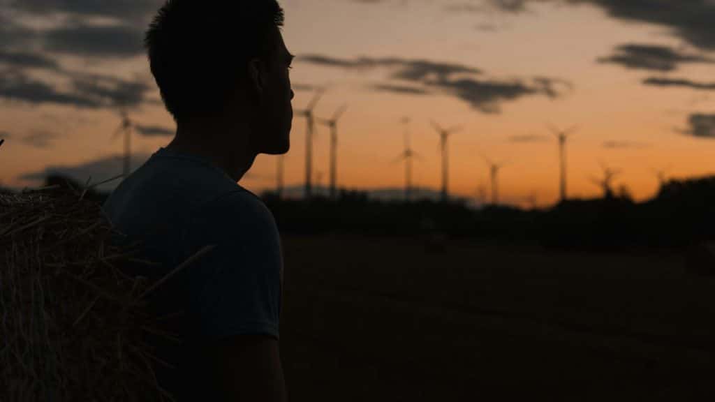 A silhouetted man standing in a field at sunset with wind turbines in the distance.