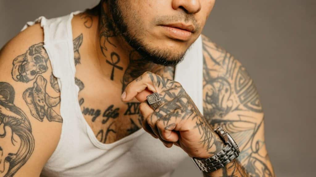 A man in a white tank top displays numerous black tattoos on his neck and arms.