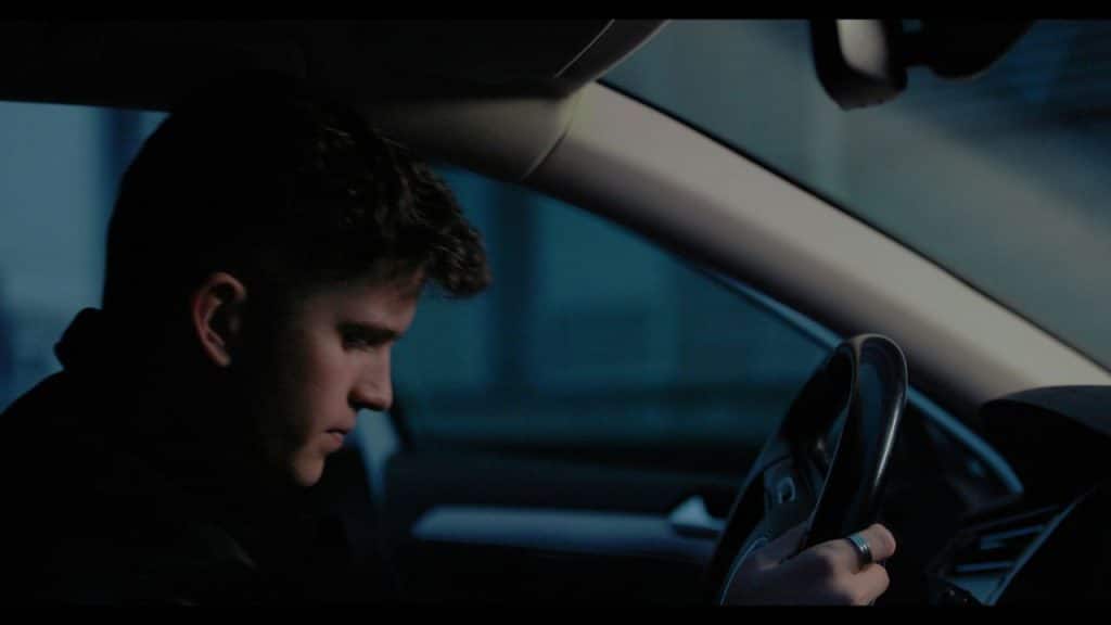 A man sits in the driver's seat of a car at night with his head tilted down.