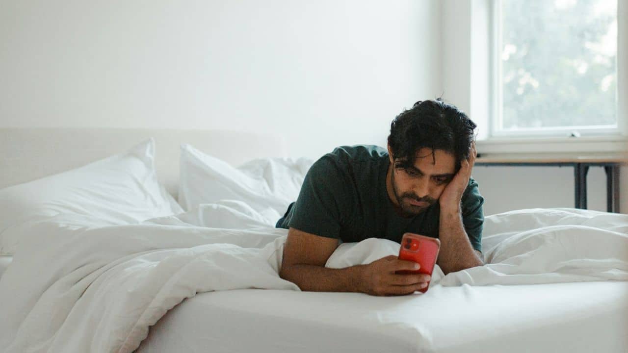A man lies in bed, propping his head up while looking at a red smartphone.