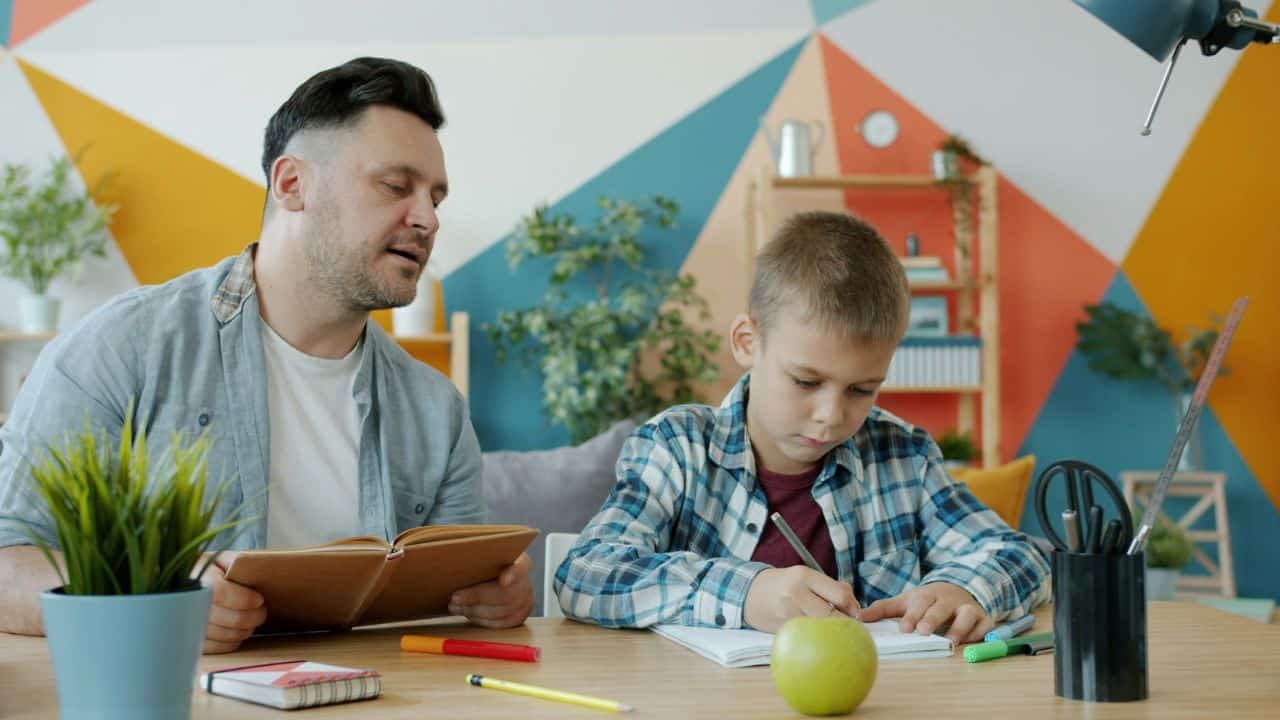 A man holds a book while a young boy writes at a table with an apple.