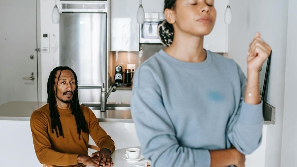 A man with dreadlocks sits at a table while a woman in a blue sweatshirt stands foreground.