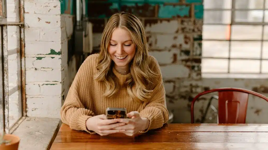 A woman smiling as she sits at a table and scrolls through her phone.