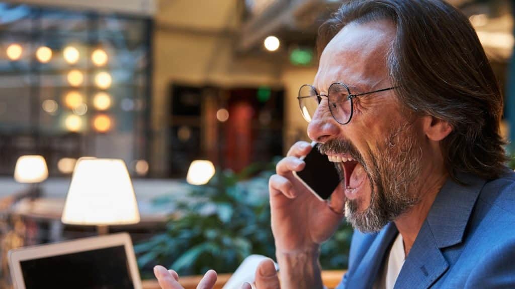 A bearded older man wearing glasses is shouting at someone over the phone.