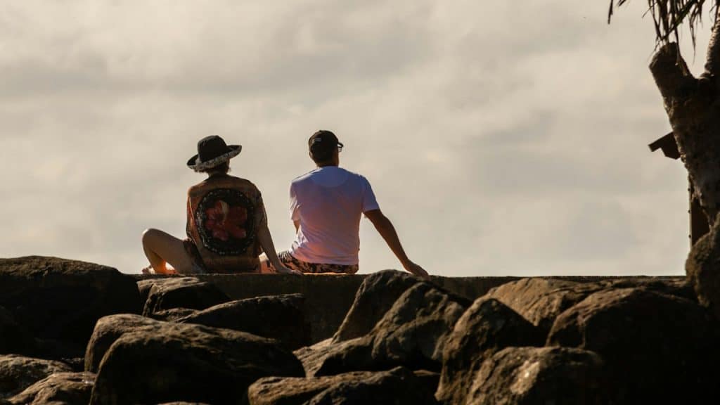 Two people sit side by side on large dark rocks, facing the open sky and horizon, with one wearing a hat and patterned shirt and the other in a light-colored T-shirt